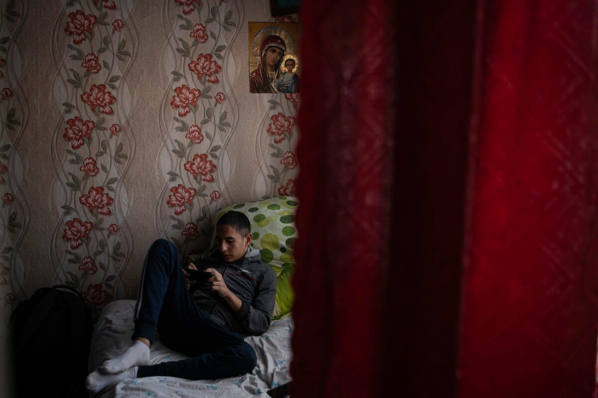 A boy sits on a bed in a room with red curtains.