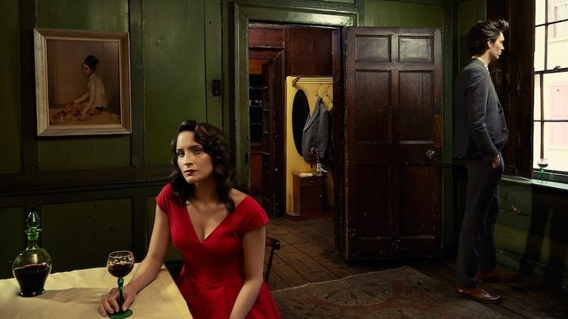 A women in a red dress with a glass of wine looks wistful at a table while behind her a man looks out a window.